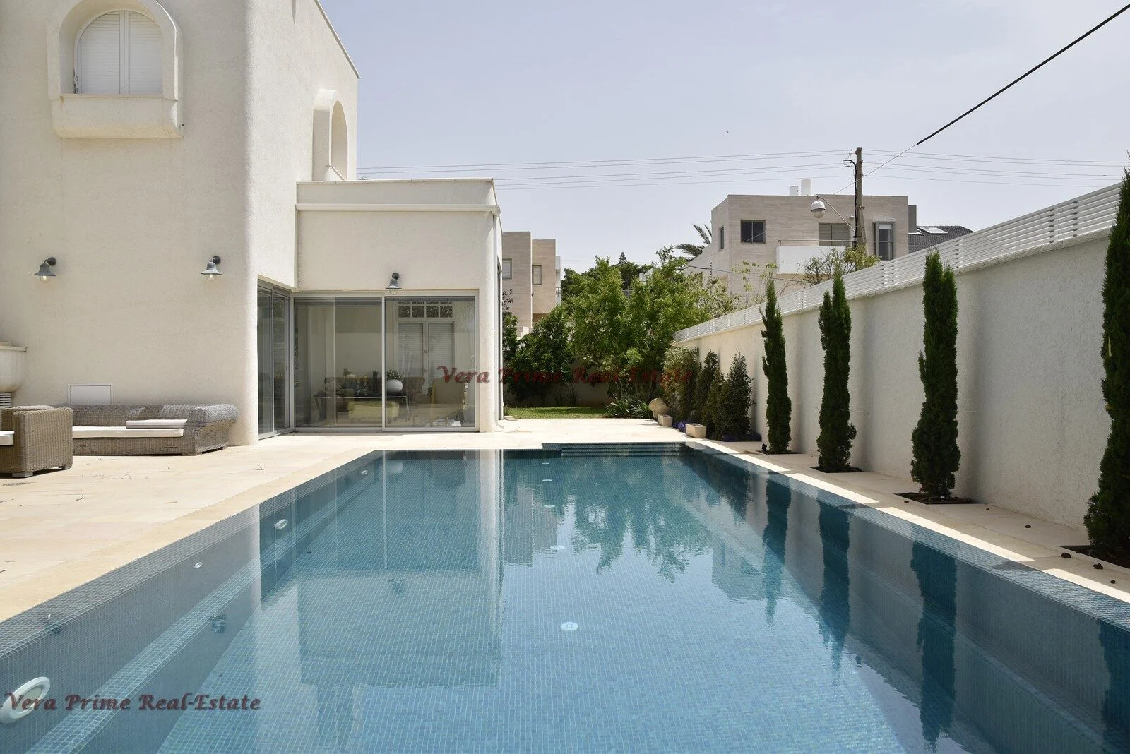 Rectangular swimming pool beside a white villa with glass doors, a paved terrace and cypress trees along a garden wall