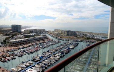 View from a balcony over a crowded marina with moored boats, waterfront buildings and the sea under a cloudy sky