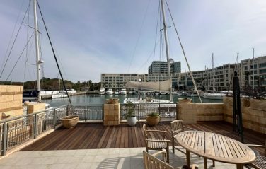 Wooden terrace with round table and chairs overlooking a marina with moored sailboats and waterfront buildings